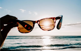 holding up designer sunglasses to see the beach and sky through, optometry near me, optometrist, eye doctor Metairie, eye doctor New Orleans, glasses store near me, eye clinic near me